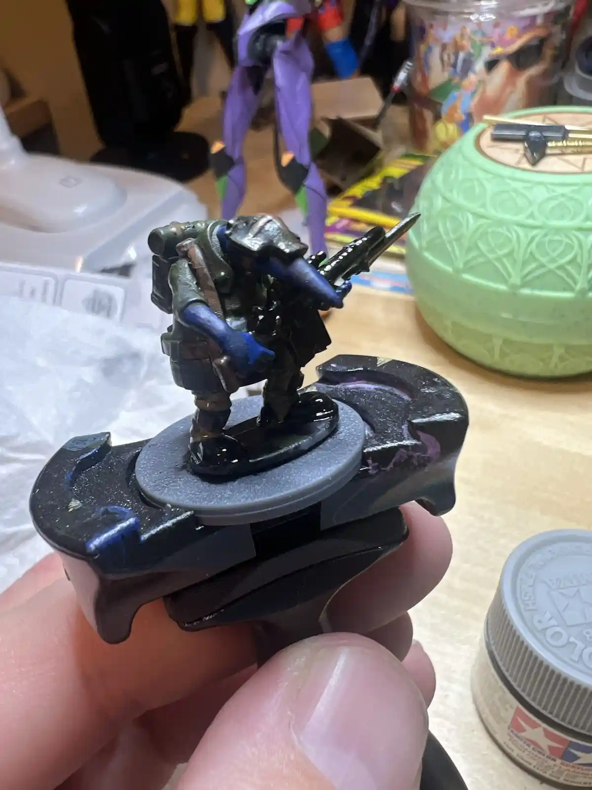 A hand holds a painted miniature soldier figurine on a black plastic base, showing blue-painted arms and a partially painted…