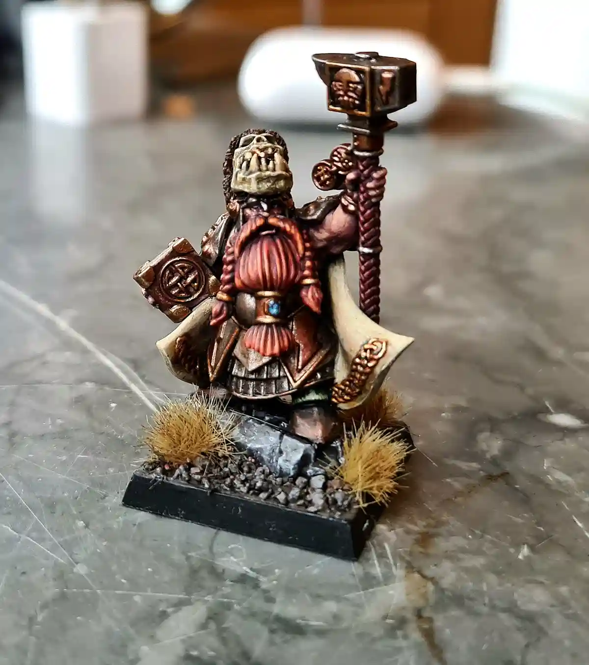 A painted miniature of a fantasy dwarf warrior with a skull helmet, wielding a large hammer and shield, standing on a rocky…