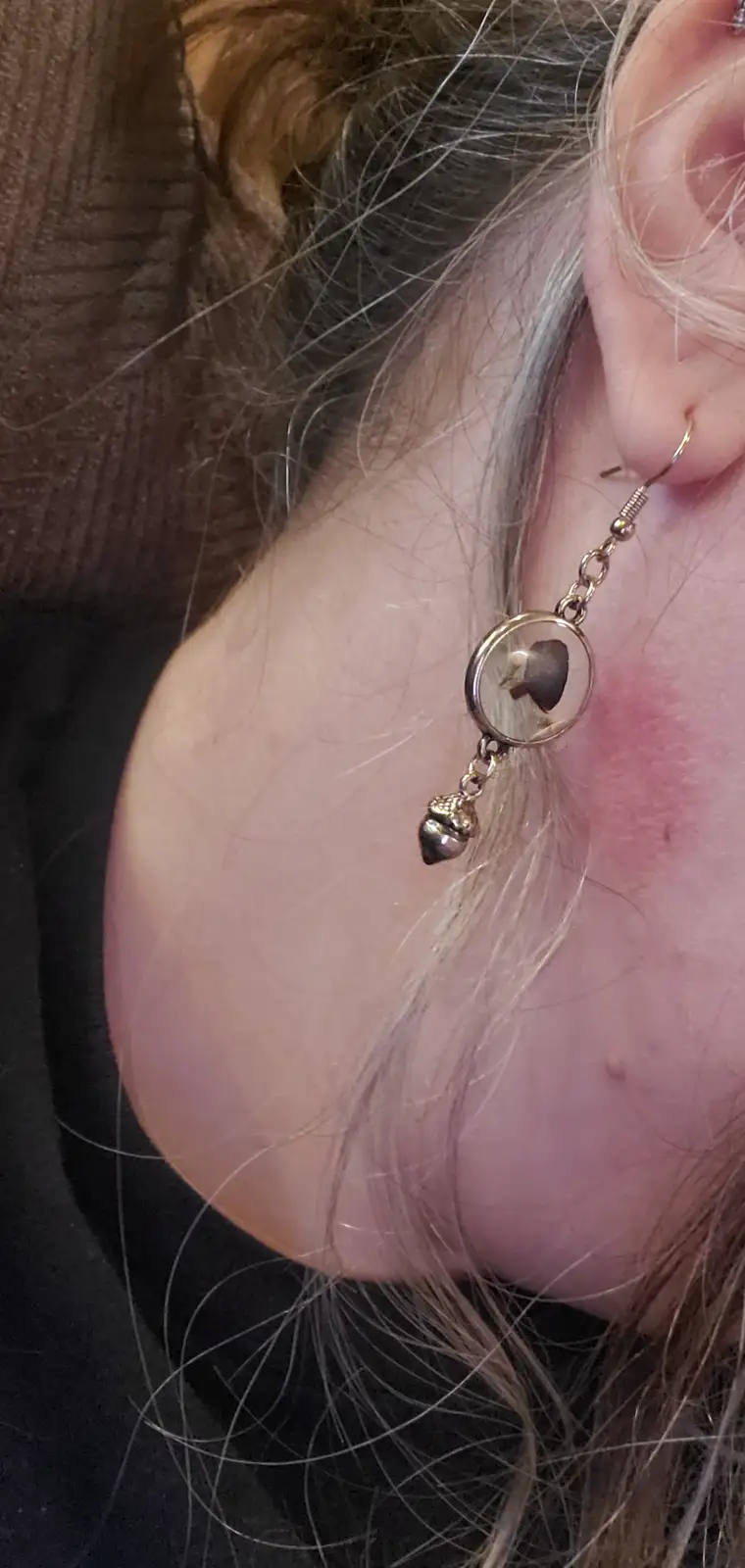 A close-up photo shows a person's ear wearing a dangling earring with a small acorn charm and a circular pendant containing…