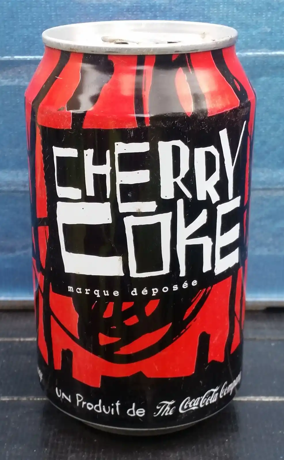 A close-up photo of a used Cherry Coke can with red and black graphics, showing the text 'CHERRY COKE', 'marque déposée'…