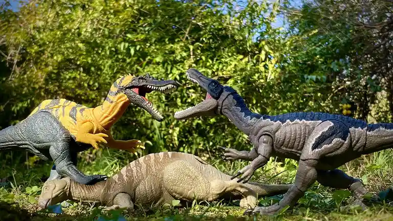 Two plastic dinosaur action figures, one yellow and black, the other gray and blue, are posed in a confrontational stance…