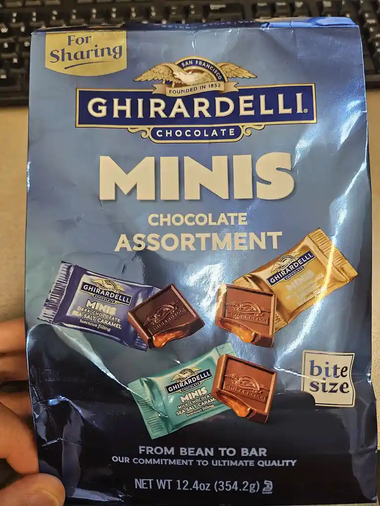 A hand holds a blue Ghirardelli Minis Chocolate Assortment bag, showing bite-sized chocolates in dark, milk, and white…