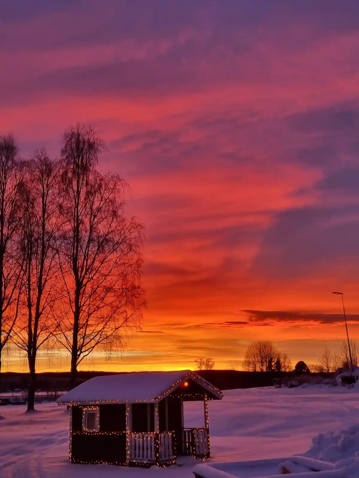 A small, snow-covered wooden cabin adorned with string lights sits in a snowy field under a vibrant, fiery sunset sky with…