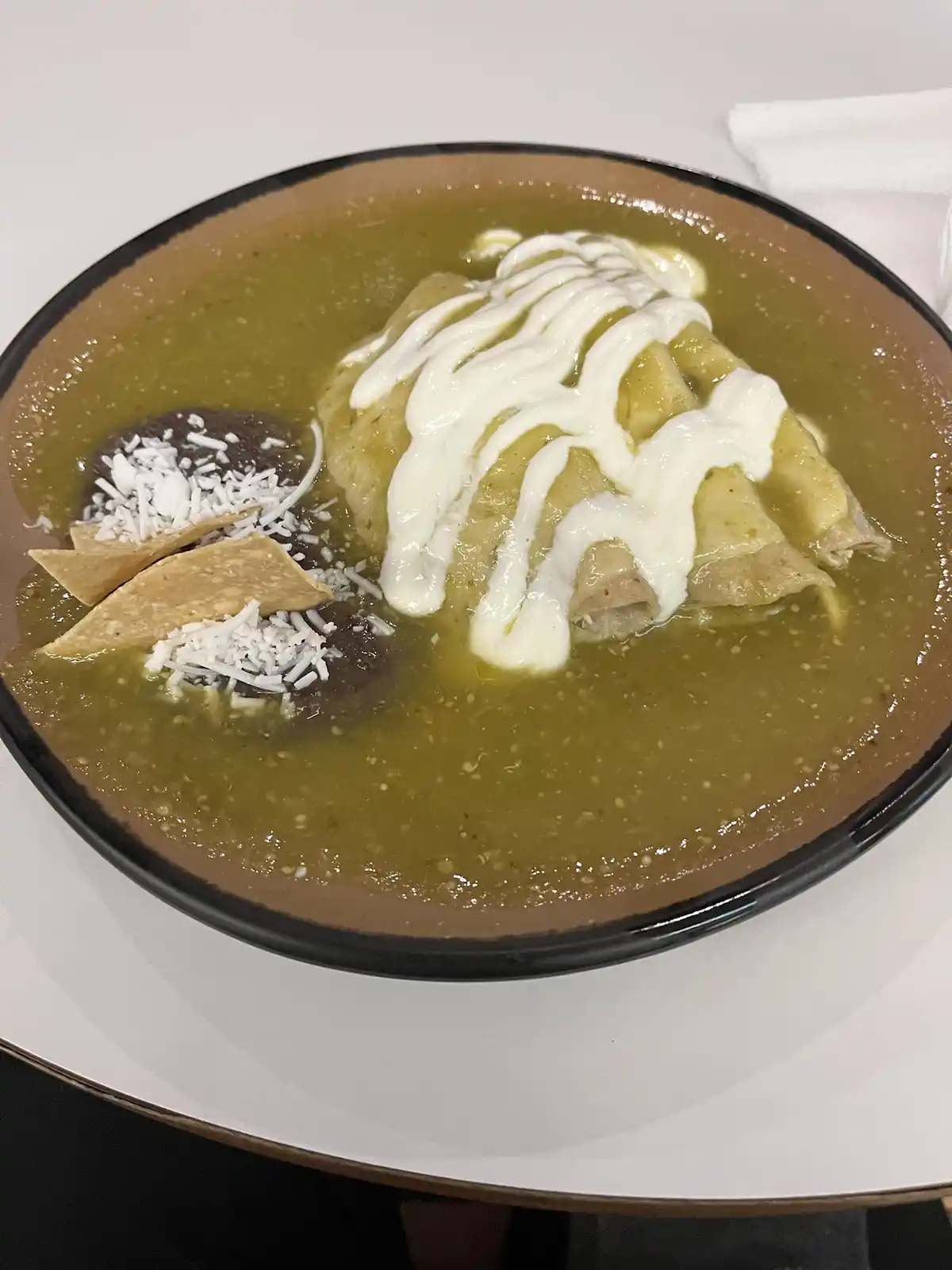 A close-up photo shows a brown bowl filled with a green, thick sauce, containing rolled enchiladas covered in white cream…