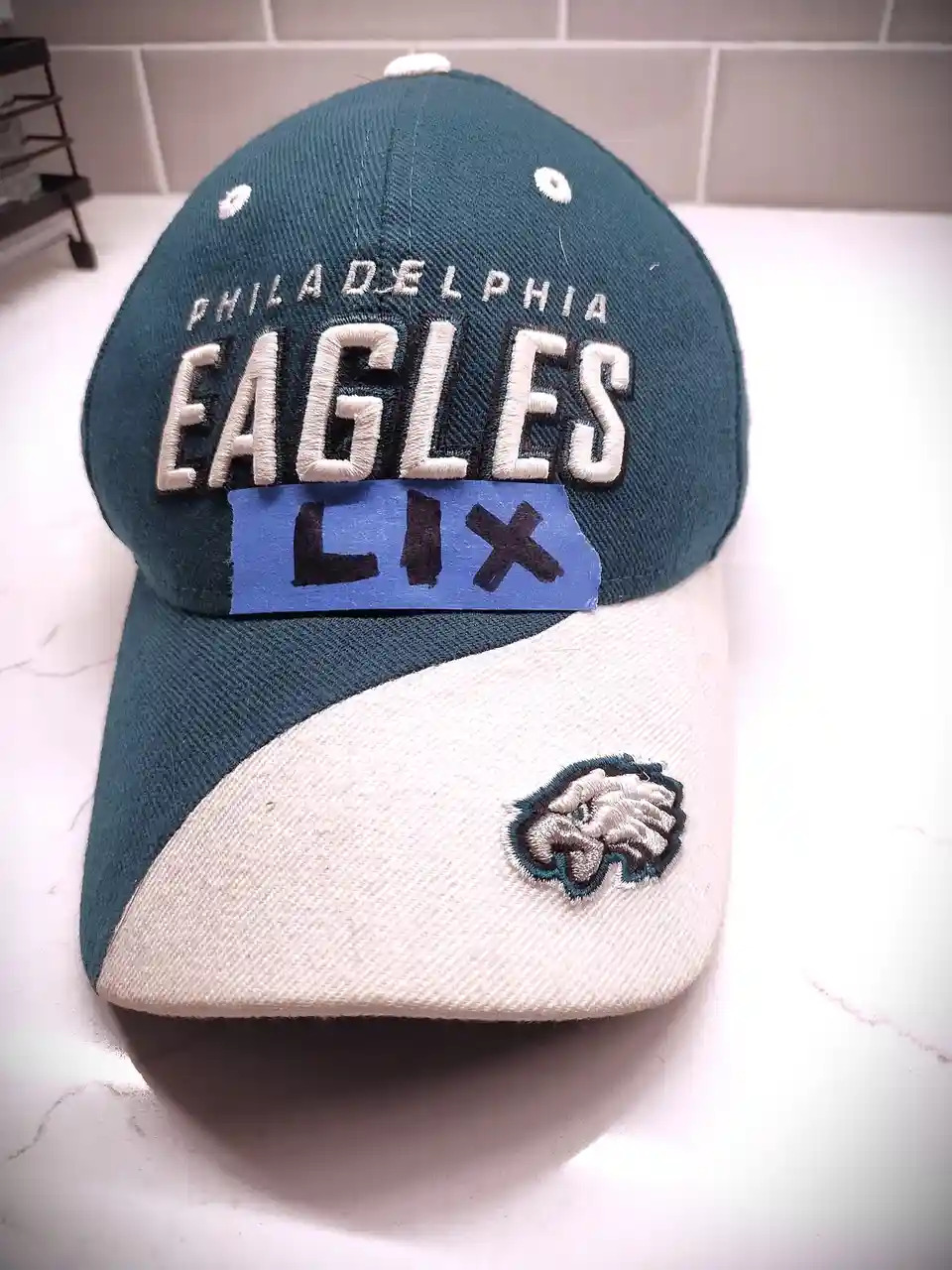 A Philadelphia Eagles baseball cap with a blue sticker over the team name that reads 'LIX' in black marker. The cap is teal…