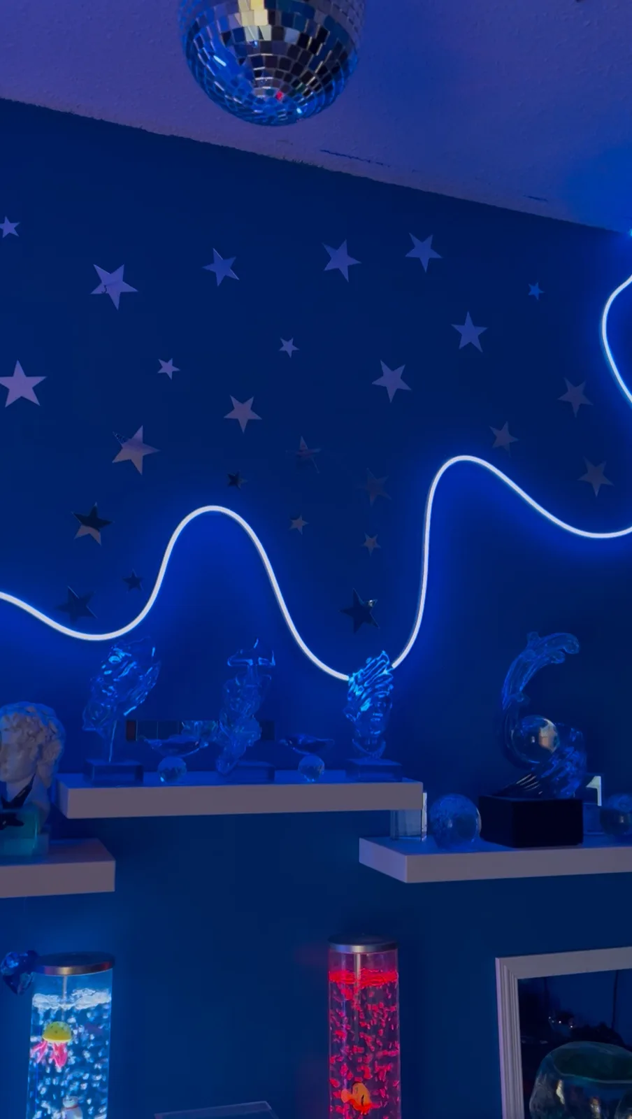 A dimly lit room with deep blue walls covered in silver star decals and a glowing white neon wave. A disco ball hangs from…