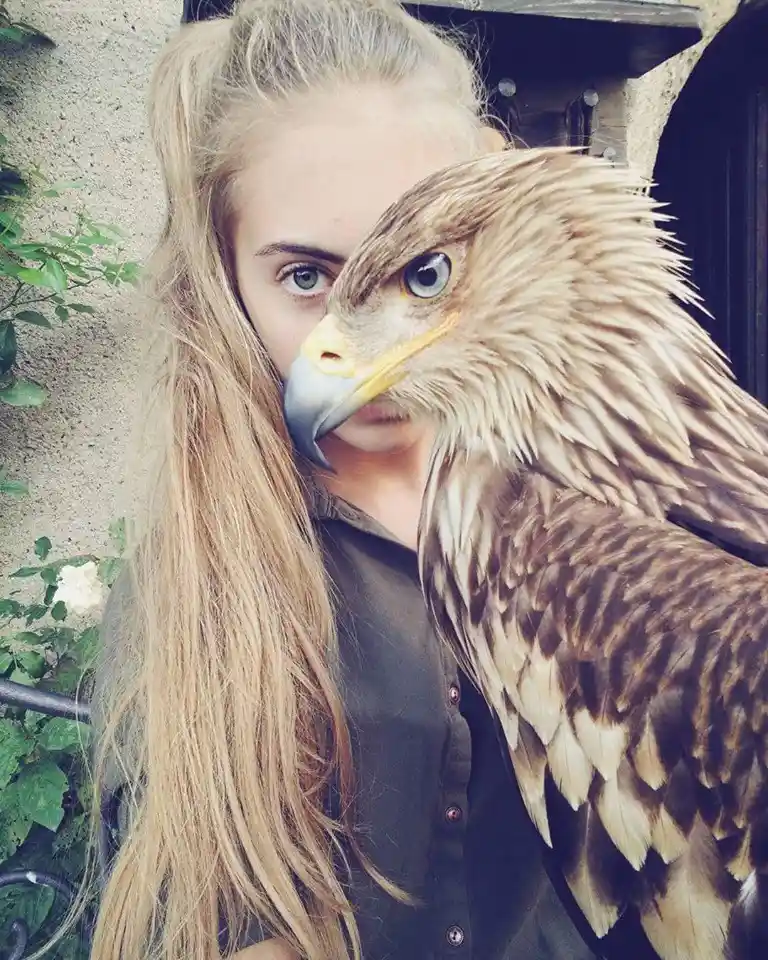 A young woman with long blonde hair and green eyes is holding a large bird of prey, likely an eagle, whose head and beak are…