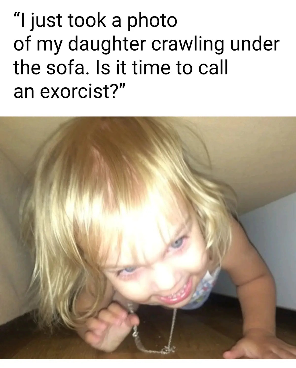 A close-up photo shows a young child with blonde hair and blue eyes crawling under a sofa, looking up with a mischievous…