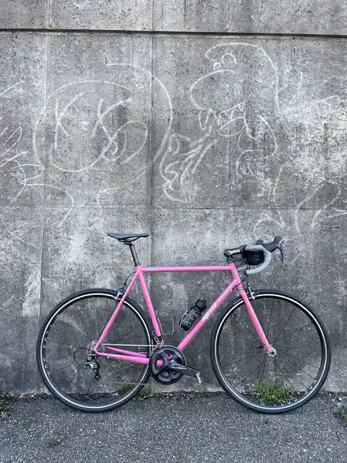 A bright pink road bicycle is parked against a gray concrete wall covered in white chalk drawings of cartoon characters. The…
