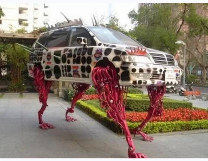 a silver suv is modified with four long, red, articulated mechanical legs, standing upright on a paved sidewalk next to a…