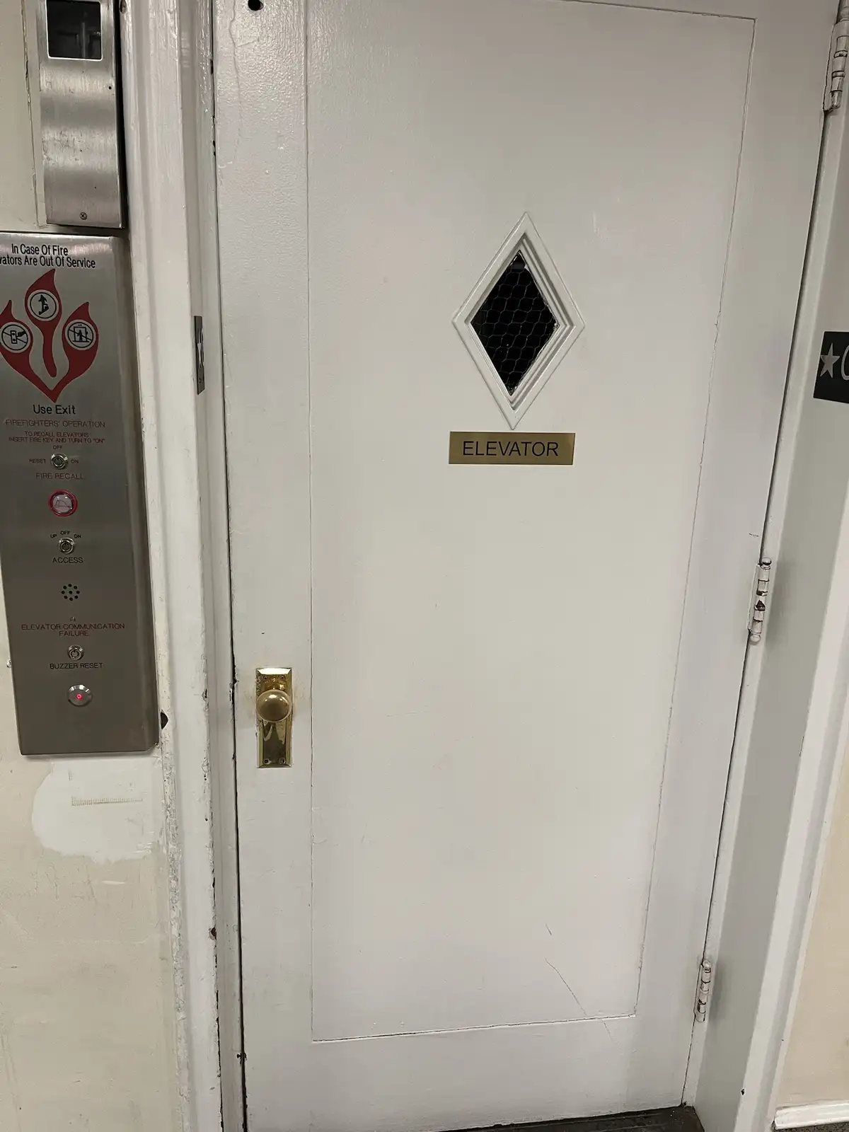A plain white door with a brass doorknob and a small diamond-shaped window with a mesh screen. Below the window is a brass…