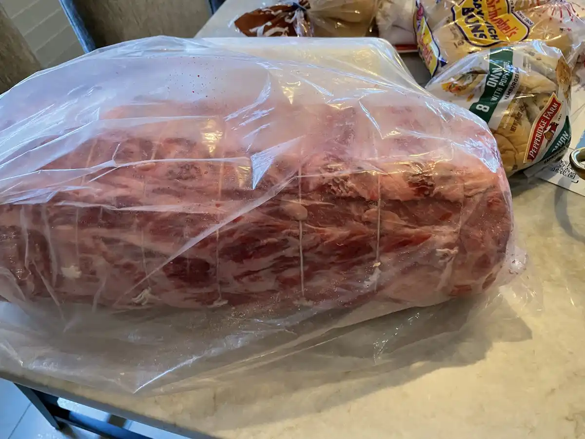 A large, raw, bone-in pork roast is wrapped in clear plastic and sits on a kitchen counter, next to a bag of Pepperidge Farm…