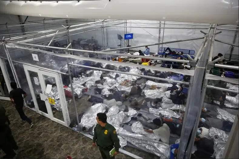An overhead view shows a large transparent tent structure filled with numerous people lying on the floor, covered with…